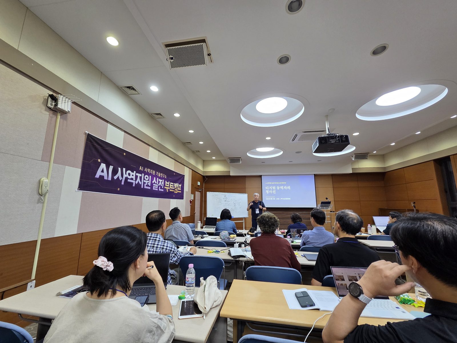 강의실 뒤쪽에서 본 모습. 벽에 '｜AI 사역지원 실전 부트캠프' 현수막이 걸려 있고, 참여자들이 책상마다 노트북을 펴 놓고 앞을 보고 있다.